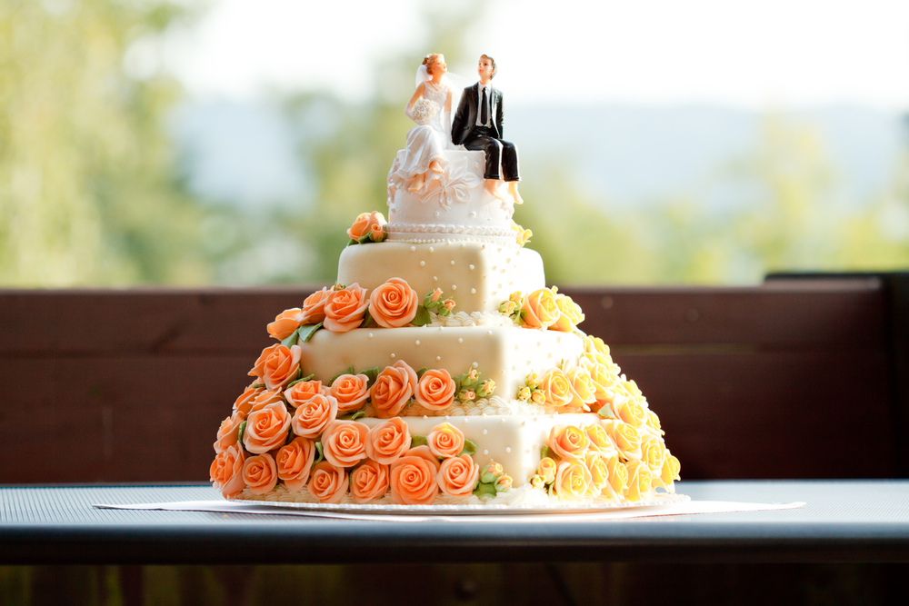 four layered square wedding cake with bride and groom figurines at the top and yellow and orange flowers on sides