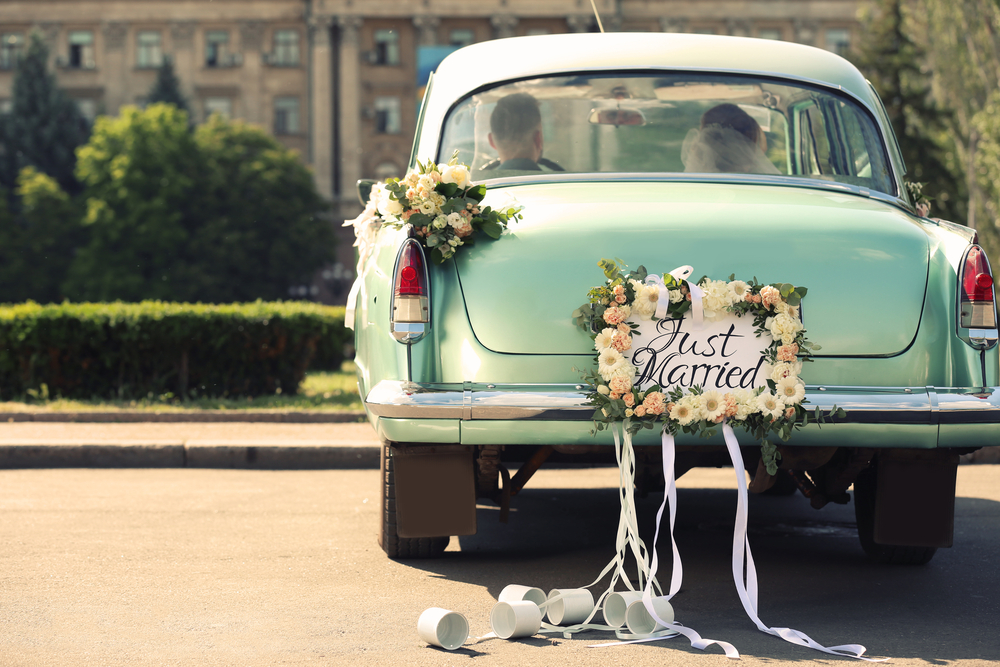 Wedding couple in car decorated with flowers, plate JUST MARRIED and cans outdoors