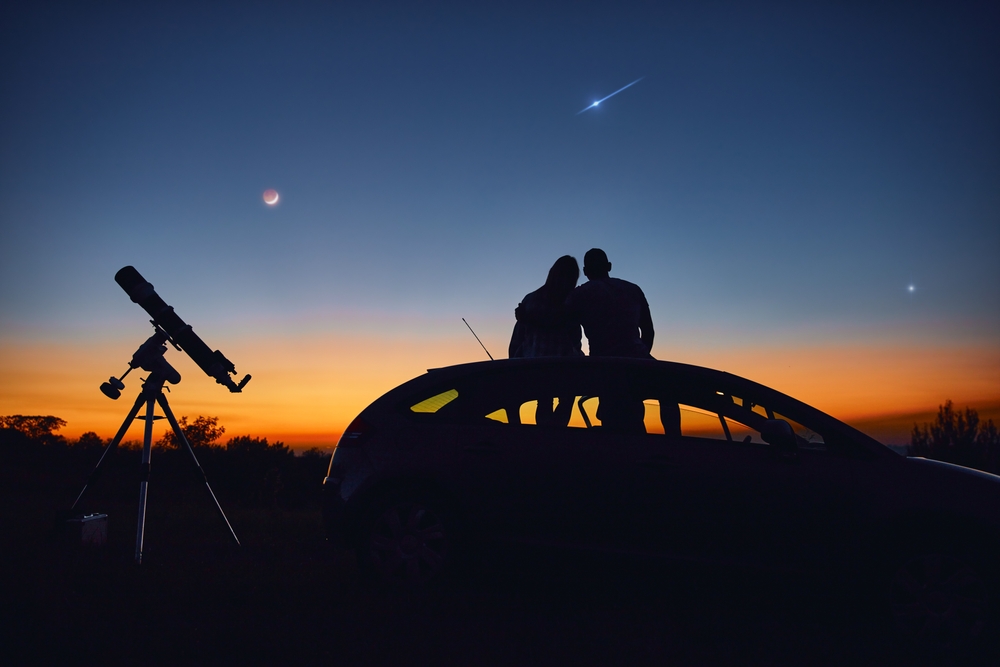 a couple on top of a car and stargazing together with an astronomical telescope