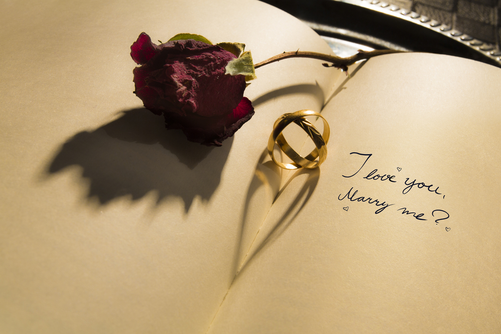 a red rose and two gold rings on top of open notebook with I Love You, Marry Me question on valentine's day