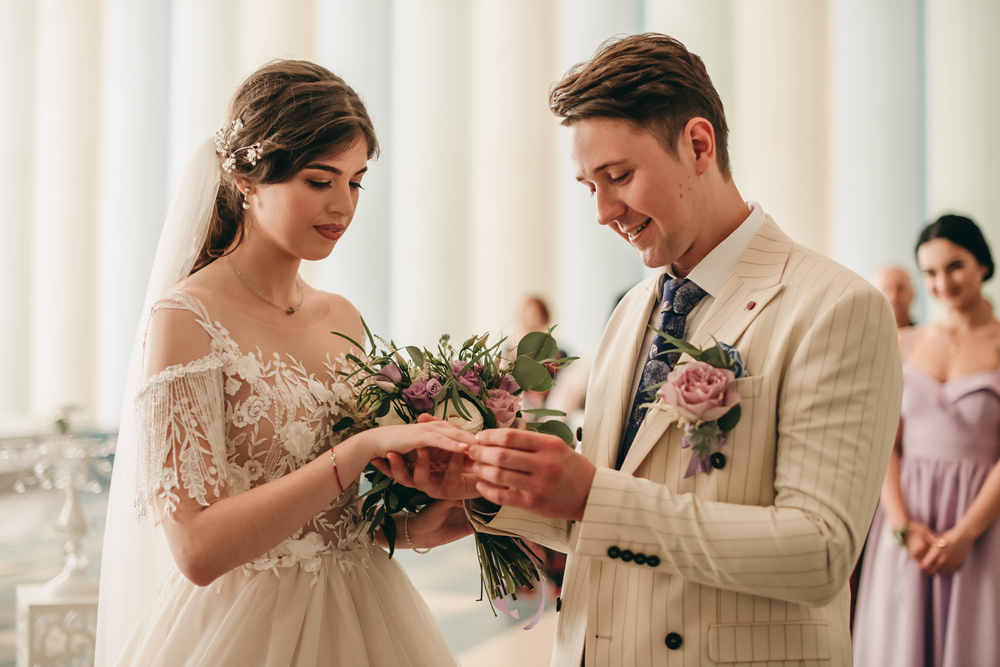 bride holding a flower bouquet and groom putting the wedding ring on bride's finger