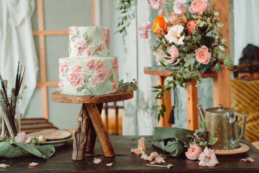 floral painted cake on wooden stand with flowers and paint tubes