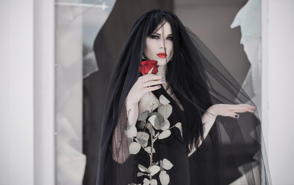 Gothic bride in black dress with veil holding a red rose and looking from a window