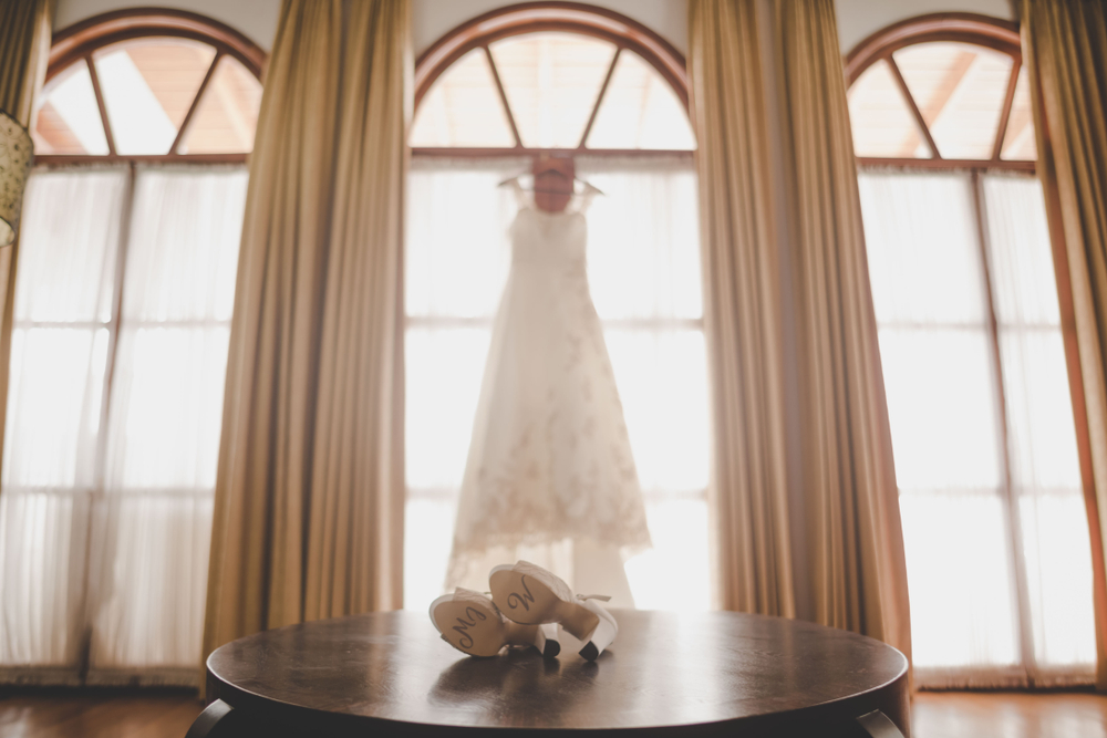 wedding dress hangin on a window and wedding shoes on round table