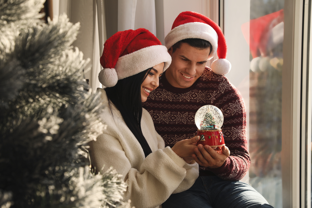 Couple in Santa hats holding a snow globe near window