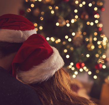 back view of a couple sitting next to a Christmas tree and wearing Santa's hats