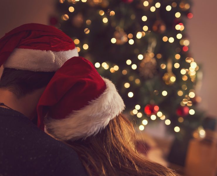 back view of a couple sitting next to a Christmas tree and wearing Santa's hats