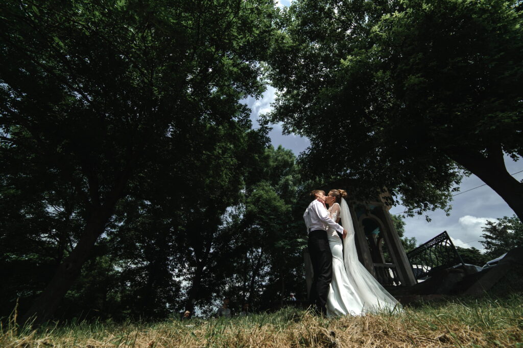 Bride and groom kissing outdoors beneath large trees near a gazebo