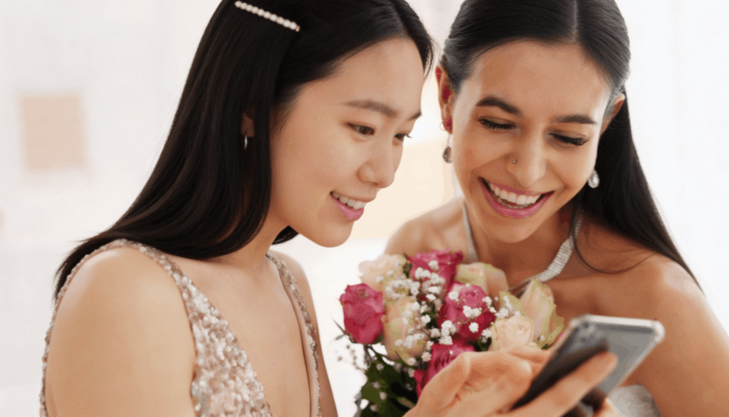 Two smiling women in formal attire look at a smartphone screen while the woman in the bridal gown holds a bouquet of pink and white roses.