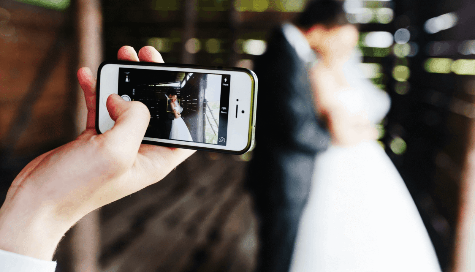 A person holds up a smartphone to photograph a bride and groom posing in a rustic wooden structure.