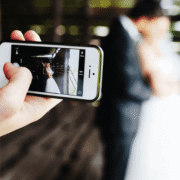 A person holds up a smartphone to photograph a bride and groom posing in a rustic wooden structure.