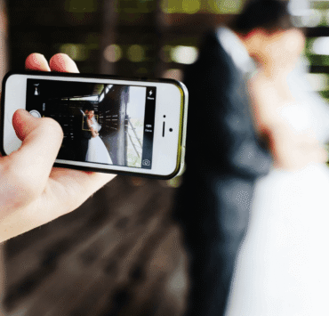 A person holds up a smartphone to photograph a bride and groom posing in a rustic wooden structure.