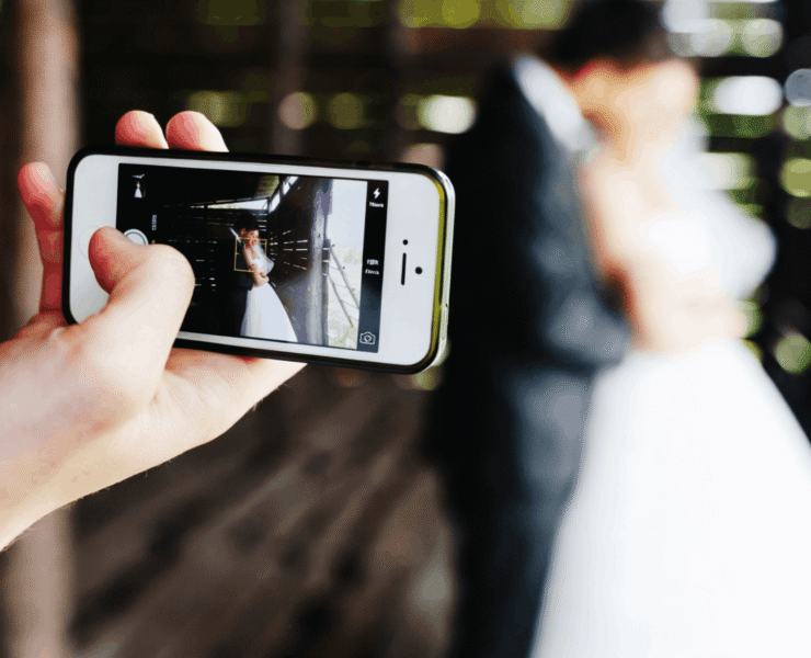 A person holds up a smartphone to photograph a bride and groom posing in a rustic wooden structure.