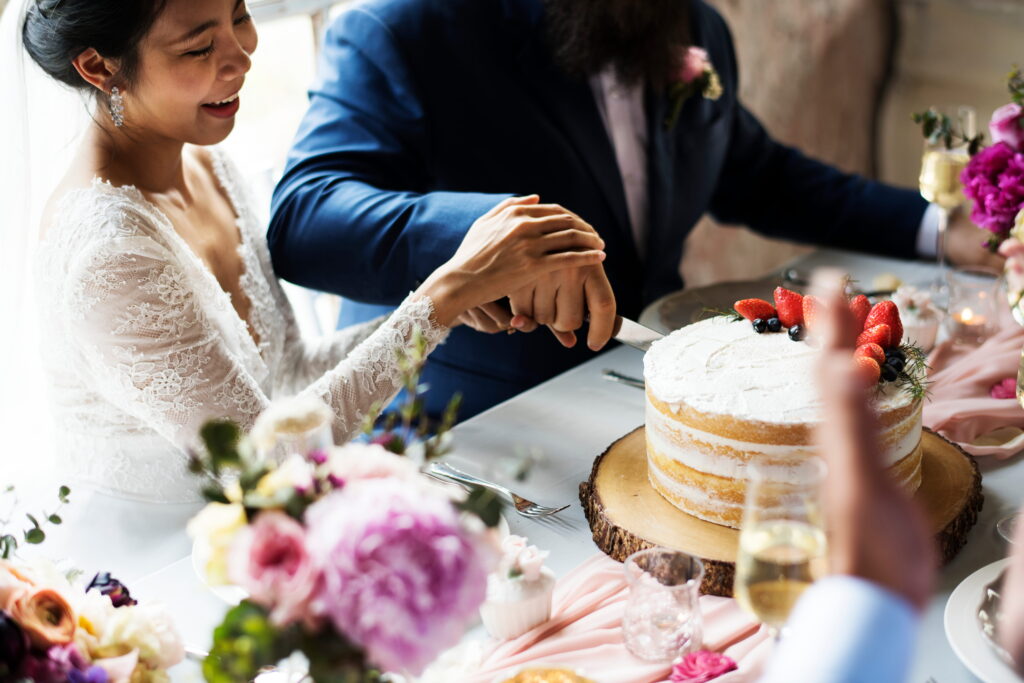 Close-up of a bride and groom's hands together using a silver knife to cut their multi-tiered white wedding cake.