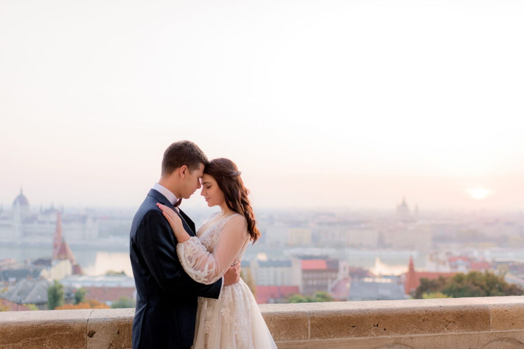 A bride and groom embrace on a balcony overlooking a scenic city skyline at sunset.