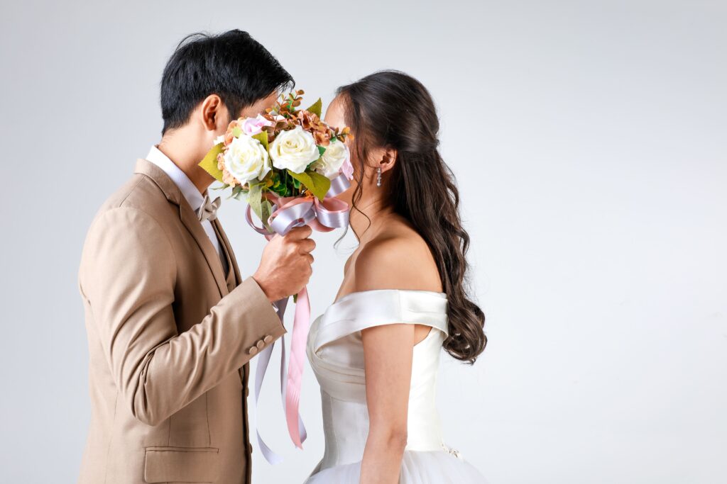 Bride and groom facing each other, faces hidden behind a bouquet of white roses with pink ribbon