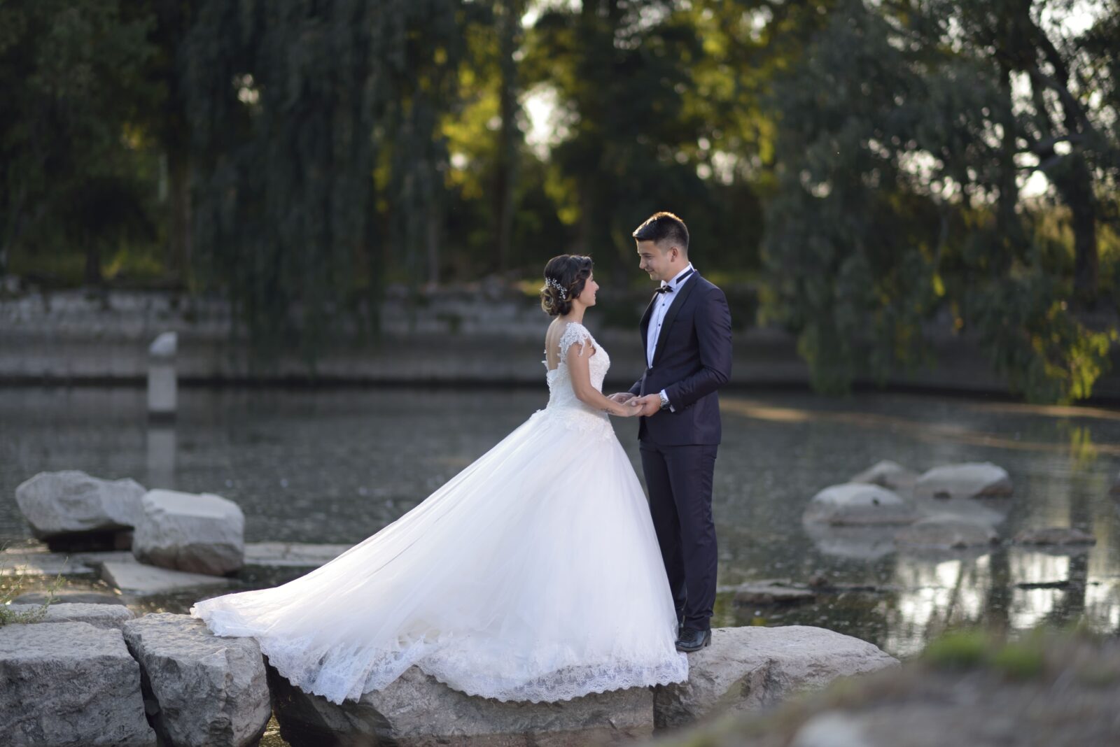 Bride in a full ball gown and groom in a dark suit holding hands on rocks beside a lake