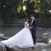 Bride in a full ball gown and groom in a dark suit holding hands on rocks beside a lake