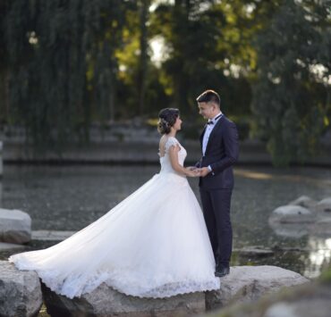 Bride in a full ball gown and groom in a dark suit holding hands on rocks beside a lake