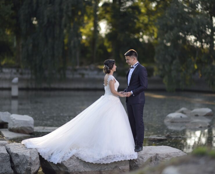 Bride in a full ball gown and groom in a dark suit holding hands on rocks beside a lake