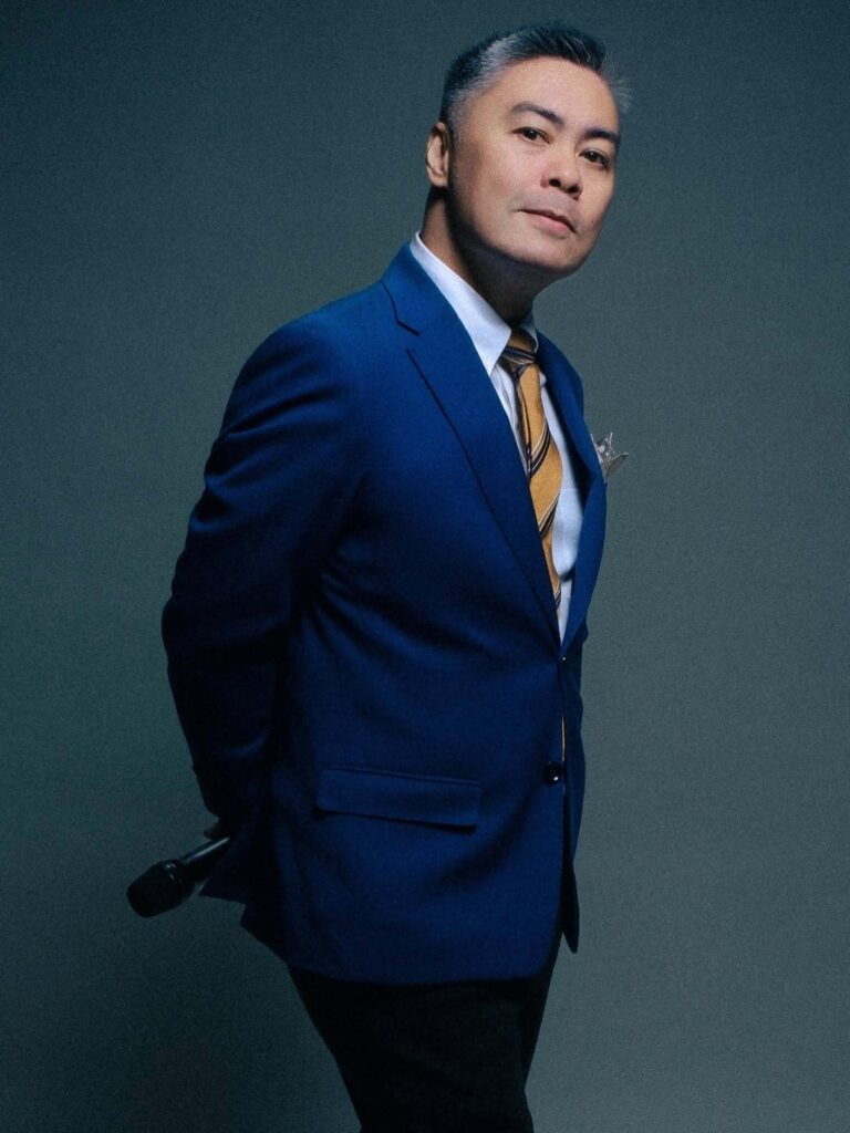 Portrait of Mike Acuña in a royal blue suit holding a microphone behind his back against a dark studio background.