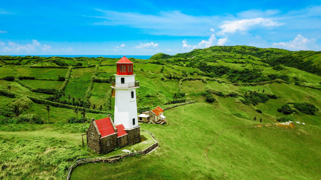 A white lighthouse with a red roof and cap standing on rolling green hills in Batanes, Philippines, with stone structures nearby and the blue ocean visible in the distance
