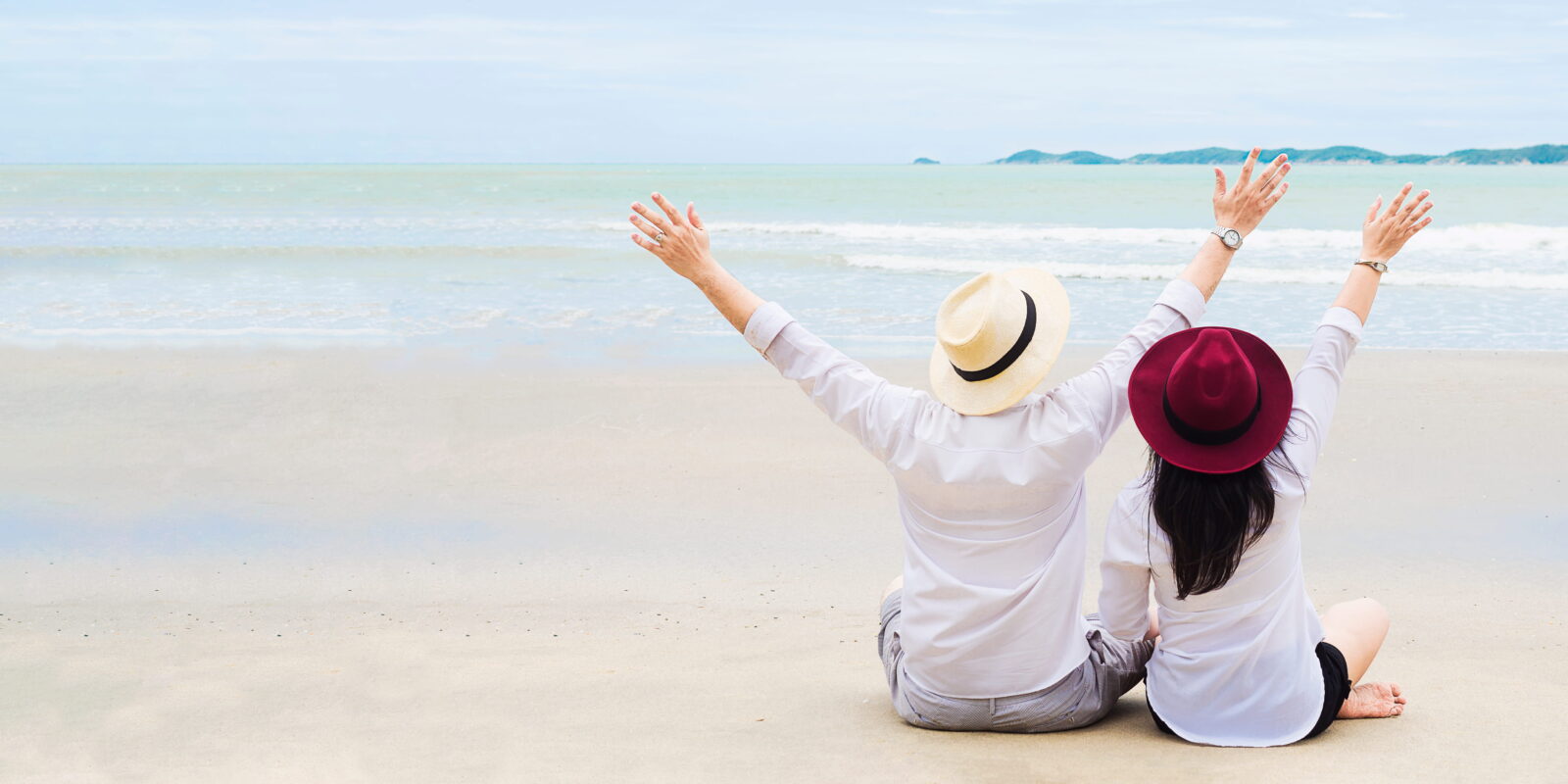 A couple sitting on a white sandy beach with their arms raised joyfully, seen from behind, facing calm ocean waters under an overcast sky