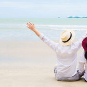 A couple sitting on a white sandy beach with their arms raised joyfully, seen from behind, facing calm ocean waters under an overcast sky