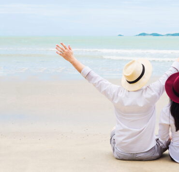 A couple sitting on a white sandy beach with their arms raised joyfully, seen from behind, facing calm ocean waters under an overcast sky