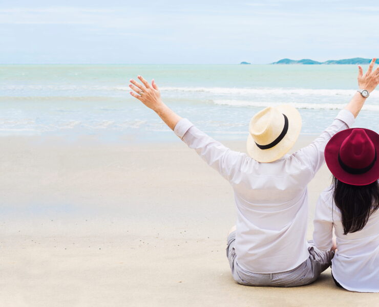 A couple sitting on a white sandy beach with their arms raised joyfully, seen from behind, facing calm ocean waters under an overcast sky