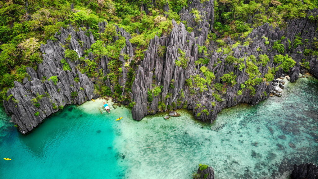 Aerial view of jagged limestone karst formations in El Nido, Palawan, surrounding crystal-clear turquoise lagoons with a traditional bangka boat and kayakers near a secluded shoreline